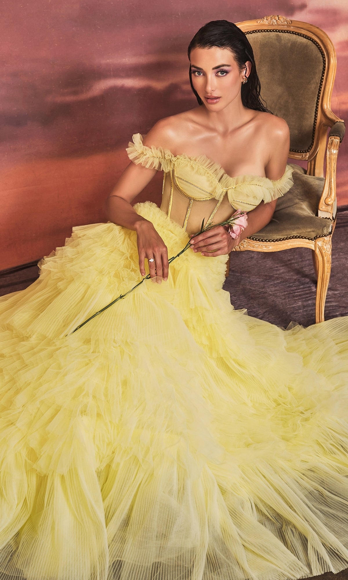 A woman in the Pastel Off-the-Shoulder Ruffled Ball Gown A1150 sits on a vintage armchair, holding a long-stemmed flower, set against a pink and brown backdrop.